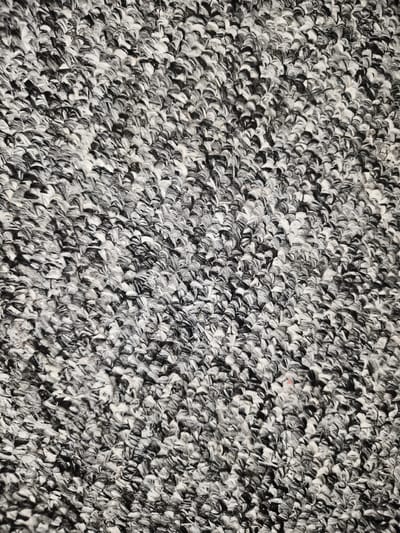 The image shows a close-up, overhead view of a textured surface, likely a carpet or rug. The texture is characterized by dense, looped fibers, predominantly in black and white, creating a mottled or marbled pattern. The fibers appear to be tightly woven and somewhat coarse. There is a small, barely perceptible reddish-pink speck on the surface, possibly a stray thread or a stain. The lighting appears to be from above, casting subtle shadows that highlight the three-dimensional texture of the material. No specific urban or natural environment can be identified from this image alone. This image does not contain any information that would allow for its localization in Солдатская, Russia, or any other specific city.