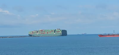 A wide-angle, horizontal shot captures a vast expanse of blue ocean under a bright, partially cloudy sky. In the foreground, the calm sea extends to the horizon. A breakwater made of large stones is visible along the left side of the frame, extending into the water.
Dominating the scene is a colossal green container ship, its deck stacked high with numerous green and red shipping containers, moving from left to right. Its immense size is emphasized by the surrounding water.
In the middle distance, several other vessels are visible. To the right of the container ship, a smaller, lighter-colored ship is seen. Further to the right, a large, red tanker ship with a white superstructure and a distinctive crane structure on its deck is moving from left to right. In the far distance, on the horizon, two more ships are faintly visible, one appearing red and another lighter in color.
The image was taken from a vantage point at sea, likely from another vessel or a coastal structure, offering a clear view of maritime activity. The general location is identified as the entrance to the Panama Canal in Cristóbal, Panama, suggested by the presence of large cargo ships and the breakwater consistent with port infrastructure. There are no people or animals visible in the frame. The overall impression is one of active international shipping and commerce.