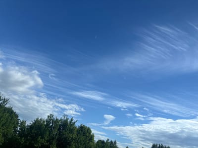 A wide-angle photograph captures a bright, clear blue sky with scattered wispy clouds and some larger, puffy clouds. A thin, crescent-shaped object, possibly a contrail or a distant celestial body, is visible in the upper center of the frame. The bottom portion of the image features the tops of dark green, leafy trees, suggesting an outdoor setting, likely in a rural or suburban area. Power lines are faintly visible stretching horizontally across the sky above the trees on the left side of the image. The overall impression is of a pleasant, sunny day. The location is Грозаўскі сельскі Савет, Belarus.