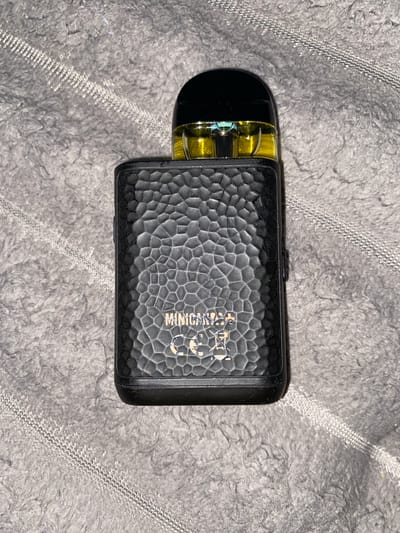 A black vape pen with a textured surface and a yellow liquid-filled cartridge is lying on a gray, fuzzy blanket. The vape pen has the text "MINICAN+" and a symbol that appears to be "CE" with a crossed-out battery underneath. The background is a soft, gray textured surface that resembles a blanket or rug. The lighting appears to be artificial, casting a slight shadow from the vape pen. There are no people or animals present in the image. The scene is static and appears to be indoors. There is no information in the image to suggest it is located in Fălești, Moldova.