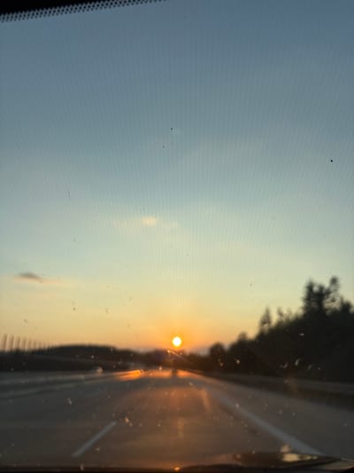 The image, taken from inside a vehicle, captures a sunset over a highway in Schnelldorf, Germany. The sky transitions from a pale blue at the top to shades of yellow and orange near the horizon. A bright, round sun is visible, positioned slightly to the left of the center of the image. The highway stretches forward, with blurred lane markings indicating movement. Trees and a dark treeline are silhouetted against the bright sky on the right side of the frame. The foreground is dominated by the vehicle's dashboard and windshield, which has some water droplets and dust specks, slightly obscuring the view. The overall impression is of a serene and picturesque drive as daylight fades.