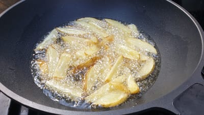The image shows potato wedges frying in hot oil in a dark, non-stick pan. The potato wedges are cut into thick, irregular shapes and are submerged in bubbling, clear oil. Some of the wedges have started to turn golden brown. The pan is set on a dark surface, possibly a stovetop or outdoor grill, with a portion of a dark, textured handle visible in the upper left corner. The scene appears to be outdoors due to the bright, natural lighting and the presence of water droplets on the rim of the pan, suggesting condensation from a cool surface or a change in temperature. The overall impression is a close-up, high-angle view of food being prepared. No people or text are visible in the image.