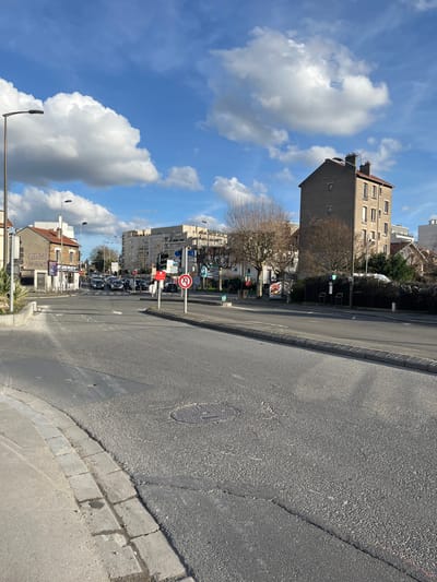 This image depicts an urban intersection in Rosny-sous-Bois, France, specifically at the confluence of Rue du 11 Novembre 1918, Avenue Jean Jaurès, and Place de l'Église. The view is oriented northeast under a clear blue sky with scattered white cumulus clouds.
The foreground consists of an asphalt road surface exhibiting cracks and patches, with a visible circular metal manhole cover. A concrete curb marks the edge of the road on the left.
To the left, a two-story building with a light-colored facade and a red-tiled roof is partially visible, housing a ground-floor establishment displaying "LEROY" and "NERGY" signage. Beyond this building, a multi-lane roadway extends into the distance, occupied by numerous stationary vehicles, primarily passenger cars, indicative of queued traffic.
The mid-ground features a traffic island or median strip with bare deciduous trees and various street signs, including a prominent red circular "no U-turn" sign. Several traffic light poles are present; one displays a green light, while another shows a red signal controlling a left-turn lane.
To the right, a distinctive, narrow, five-story residential building constructed with grey brick and topped with a red-tiled roof and two chimneys is prominent. The right side of the roadway features a sidewalk with bare trees, green shrubbery, bicycle racks, and a bus stop shelter. In the background, additional multi-story urban buildings are visible. No individuals are discernibly present within the frame.