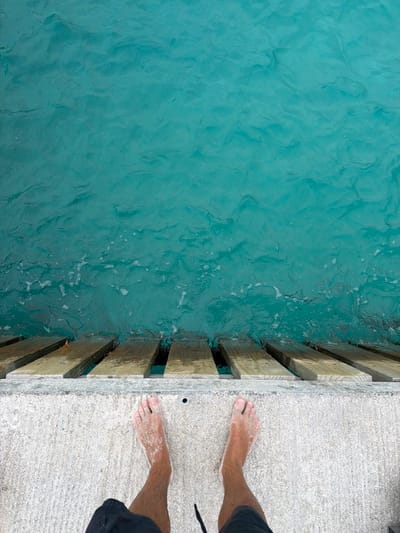 Captured from an overhead perspective, the image presents a concrete pier situated at Fulidhoo, Maldives, abutting the turquoise sea. A portion of a person is visible, with their sandy feet resting on the concrete pier near the wooden planks separating the pier from the water. The sea water displays a rippled surface, contributing to the overall serene atmosphere of the scene.