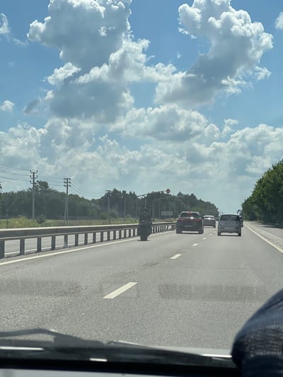A black motorcycle with a rider is traveling on a highway in Novomyshastovskaya, Russia, under a partly cloudy sky. The motorcycle is in the left lane, and several cars are ahead of it. The highway has a concrete barrier on the left and a grassy shoulder with trees and utility poles on the right. The rider is wearing a black outfit and a helmet, and the motorcycle appears to be a sportbike. The cars are also traveling in the same direction, and the scene suggests a normal day of travel on a highway.
