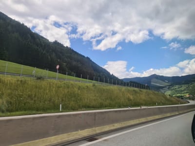 This is a photo taken from a vehicle on a highway in Unzmarkt-Frauenburg, Austria. The highway has a concrete barrier separating the lanes. On the right side of the highway, there is a steep, grassy embankment with a fence running along the top. Beyond the embankment, a lush green mountainside covered in trees dominates the landscape. The sky is partly cloudy with patches of blue. The overall scene depicts a scenic drive through a mountainous region in Austria.