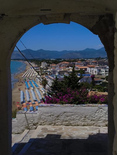 An eye-level, medium shot captures a view through an arched opening, framed by rough, textured white walls on the sides and top. The opening looks out onto a sun-drenched coastal town and beach scene under a clear blue sky. In the foreground, within the arch, is a white-washed stone railing topped with a decorative tiled border, behind which vibrant purple bougainvillea flowers bloom. Below the railing, a shadow-dappled stone staircase descends.

Beyond the immediate foreground, the scene opens to a wide, sandy beach lined with rows of blue and white beach umbrellas and lounge chairs. Numerous people are visible on the beach, some walking, others sunbathing. The turquoise sea stretches to the left, meeting the shore. Further inland, a dense collection of buildings with red-tiled roofs and white facades make up the town. In the distance, hazy blue mountains form the backdrop. The overall impression is a vibrant, populated seaside resort. A sign is visible at the top of the arch which reads "COSTA ANTONIO LAURELE."