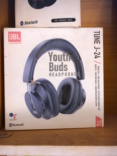 The scene depicts two commercially packaged headphone boxes resting on a light brown wooden shelf in the city of Ijara, Nigeria. The prominent foreground box is white cardboard, sealed in clear plastic, advertising "JBL Youth Buds HEADPHONES" model "TUNE J-24." An image of black over-ear headphones dominates the front. Printed features include "Wireless," "Pure Bass Sound," "Active Noise Cancelling," "Google Assistant," and "Bluetooth," alongside a "48" battery icon. The box exhibits minor surface wear. Partially visible behind it is a second white cardboard box, indicating "Bluetooth," "FM • RADIO • MP3" capabilities. No individuals or active interactions are present.
