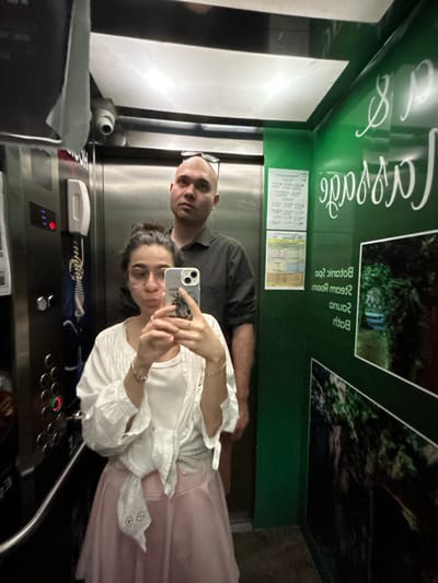 A selfie captured inside an elevator shows a woman in the foreground taking the photo with a smartphone, alongside a man standing slightly behind her. The image is rotated 90 degrees counter-clockwise. The woman, with dark hair and glasses, is wearing a white ruffled top and a light pink skirt. She has a watch or bracelet on her left wrist and rings on her fingers. The man, bald and with glasses pushed up onto his head, wears a dark grey short-sleeved shirt. Both individuals look towards the camera with neutral to slightly smiling expressions, appearing relaxed.

The elevator interior features reflective metallic panels on one side, where the individuals are visible, and dark green panels on the other side. The green panels are adorned with white, flowing script text and images depicting lush green foliage, suggesting a natural or spa-like theme. Visible text on the green wall, partially reflected, includes "Spa & Massage" (with the first word partially obscured, possibly "Caspas"), and mirrored text like "Sauna," "Steam Room," and "Botanic Spa." Two smaller, rectangular information sheets, likely containing schedules or service details, are also affixed to the green wall, though their text is illegible.

The elevator control panel is visible below the individuals, featuring an array of buttons and an illuminated red light, possibly indicating an active function or call. A white intercom handset is mounted to the left of the control panel, and a small security camera or sensor is positioned above it. The scene captures a casual moment of travel, likely within a hotel, spa, or wellness center in Esenler, Turkey, given the provided location context. The lighting is internal, so no time of day or weather details are discernible.