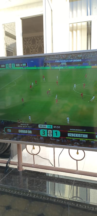 The image shows a television screen displaying a football match. The score is Congo DR 3, Uzbekistan 1. The match is part of the FIFA World Cup 2026. The screen also shows player names and timestamps, with "MAYALE 78'" and "WISSA 68' (P), 90+1'" for Congo DR, and "SHOMURODOV 10'" for Uzbekistan. The time displayed on the screen is 90:00 +8, with an additional 01:51. The scene is a close-up of the TV screen, which is placed on a stand in an outdoor setting. A metal railing is visible in the foreground, along with a dark reflective surface that appears to be a table or counter. The background shows part of a building with white walls and windows. The text mentions "Baghdad, Uzbekistan," but this is likely a misinterpretation as Baghdad is the capital of Iraq, not Uzbekistan. There is no visual evidence to confirm or deny the location being in Baghdad, Uzbekistan, as the image only shows a television screen and its immediate surroundings.