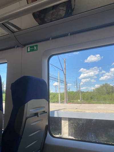 The image shows the interior of a train carriage, viewed from a passenger's seat. A dark blue seat is partially visible in the foreground, with the back of another seat in front of it, featuring a gray foldable tray. Above and to the left, a green exit sign with a white pictogram of a person running towards an arrow is affixed to the wall. The train is either at a station or moving slowly past one, as indicated by the presence of a platform with yellow markings and overhead railway electrical lines. Lush green trees and vegetation line the track. The sky is blue with scattered white clouds. The scene appears to be in a suburban or rural area, possibly on the outskirts of a city. While the specific location within Primorsky Krai, Russia, cannot be definitively identified without further context, the presence of railway infrastructure and natural scenery is characteristic of many areas within the region.