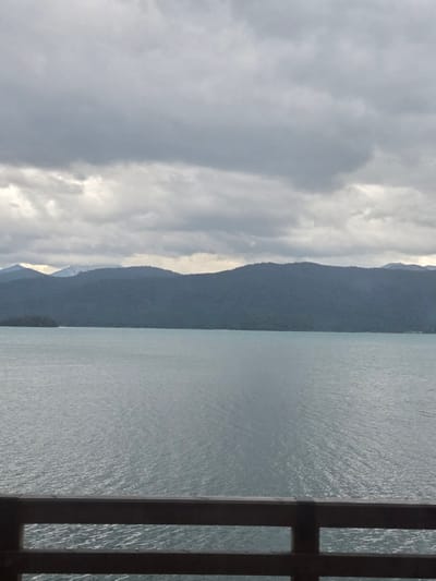 The image shows a body of water with a forested mountain range in the background under a cloudy sky. In the foreground, there is a wooden railing. The scene is overcast and appears to be a natural landscape. There are no people or discernible urban structures visible in the image. The location is not Kochel am See, Germany.