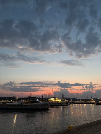 The image captures a sunset over a marina in Sopot, Poland. The sky is filled with dramatic clouds, painted in hues of gray, blue, and orange from the setting sun. Below, a calm body of water reflects the vibrant colors of the sky and the lights of the marina. Several boats, including yachts and sailboats, are docked at piers, their masts silhouetted against the colorful sky. The marina is illuminated by streetlights and lights from buildings along the waterfront, creating a warm glow that contrasts with the deepening twilight. The foreground shows a portion of a wooden pier with a yellow bollard. The scene evokes a peaceful and serene atmosphere.