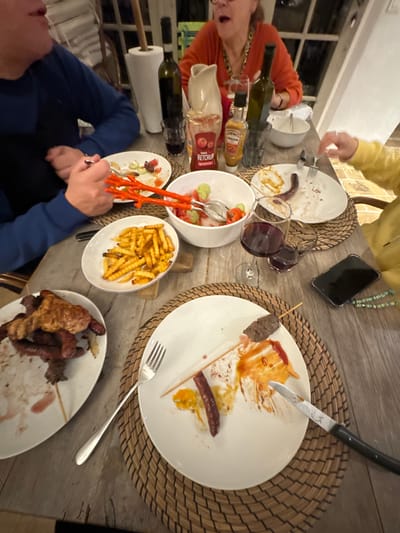 The scene depicts an indoor dinner gathering around a rustic wooden table in Tourrettes-sur-Loup, France. Four individuals are partially visible, engaged in a casual meal under warm artificial lighting.
On the left, a person wearing a blue long-sleeve top holds a silver fork over a white plate containing remnants of a salad, including olives and cucumber slices, and red sauce. To the upper center, an individual in an orange long-sleeve top with a visible gold-toned necklace has an open mouth, suggesting mid-conversation or eating. To their right, a hand wearing a yellow long-sleeve top holds a fork over a white plate containing a single sausage link and smears of red and yellow sauce. In the bottom-left, a plate holds several grilled meat items, including sausages and larger cuts, with some consumed.
The table is adorned with woven placemats under several white plates, many showing varying stages of consumption. A prominent plate in the foreground features a partially eaten meat skewer and a single sausage, surrounded by ketchup and other sauces. Beside it are a silver fork and a dark-handled knife with an engraved emblem. A white bowl filled with golden french fries is centrally placed, next to another white bowl containing a salad of sliced cucumbers and tomatoes, served with orange plastic tongs.
Beverages include a wine glass with red wine, an empty glass, and two dark bottles, likely wine or olive oil. Condiments include bottles of Heinz Ketchup and Heinz Andalouse sauce. A roll of white paper towels on a wooden holder is visible in the background. A black smartphone in a clear case with a green and white beaded strap rests on the table. The background reveals a white-framed window with multiple panes.