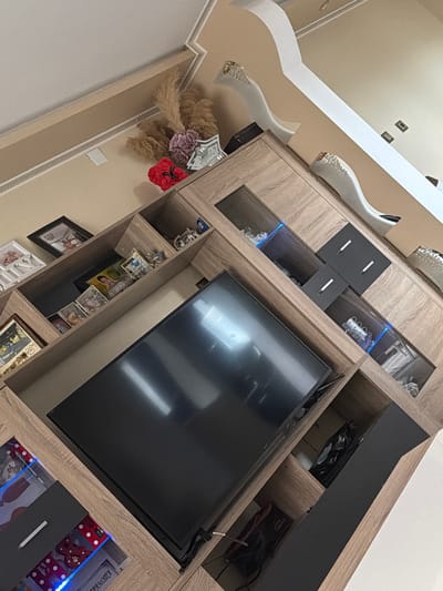 The image shows a modern living room interior. The focal point is a large, wall-mounted entertainment unit with a flat-screen television. The unit is made of wood with gray accents and includes enclosed cabinets and open shelves displaying decorative items such as vases, picture frames, and other ornaments. The ceiling features ornate, white decorative moldings. No people are visible. The location is suggested to be in Velichkovo, Bulgaria, based on the prompt, but the visual data provides no concrete evidence to confirm this location.