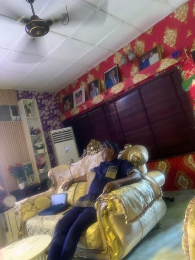 The image captures an opulent indoor scene, described as being in Akpajo, Nigeria, likely a living room. A single adult male is seated on a large, ornate gold-colored sofa upholstered with a light-colored, patterned fabric. He is dressed in a navy blue traditional outfit featuring gold embellishments and wears a matching cap. His gaze is directed towards his left. A small open laptop or tablet rests on the sofa beside his left leg.

The room's ceiling is composed of white, recessed panels, from which a black and gold ceiling fan with blurred, rotating blades is suspended. The right wall is covered in distinctive red wallpaper with prominent gold, swirling patterns, against which several framed portraits and two decorative wall sconces are mounted. A large window area on this wall is obscured by dark brown horizontal blinds. The left side of the room features a section of light brown vertical paneling adjacent to a wall adorned with purple and white geometric patterned wallpaper. A tall, white, multi-tiered shelving unit stands against this wall, displaying various decorative items, including artificial flowers and ornaments. Both a wall-mounted and a floor-standing air conditioning unit are visible on the left side of the room. A small, light-colored round coffee table is partially visible in the foreground. A pink alarm clock and another framed picture are on a surface to the far left.