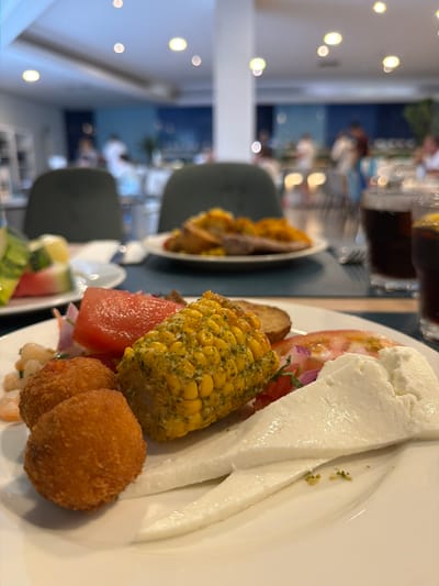 A close-up shot shows a white plate with a buffet-style meal. The plate holds fried croquettes, a slice of watermelon, a piece of seasoned corn on the cob, and slices of white cheese. In the background, other plates of food are visible on a table, along with glasses of dark liquid, possibly soda. The setting appears to be a restaurant or hotel dining area, with blurred figures of people seated at tables in the distance. The overall lighting suggests an indoor environment, possibly during daytime or evening. The image is taken in Las Dehesas, Spain.