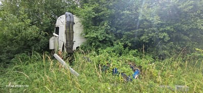 A white truck cab is overgrown with vegetation, partially hidden by trees and tall grass. The cab appears damaged, with bent metal and exposed components. A large, grey, angled object, possibly a piece of concrete or metal, lies in the foreground. In the background, blue barrels are visible among the dense foliage. The scene is set in a natural environment, likely a field or roadside, with lush green trees and plants. The lighting suggests it is daytime, with some sunlight filtering through the leaves, creating lens flare. The overall impression is one of abandonment and decay, with nature reclaiming the man-made structure. The visible text at the bottom left corner reads "01. Juli 2026 17:02", indicating the date and time the photo was taken. The location is Auerbach in der Oberpfalz, Germany. There are no people visible in the image.