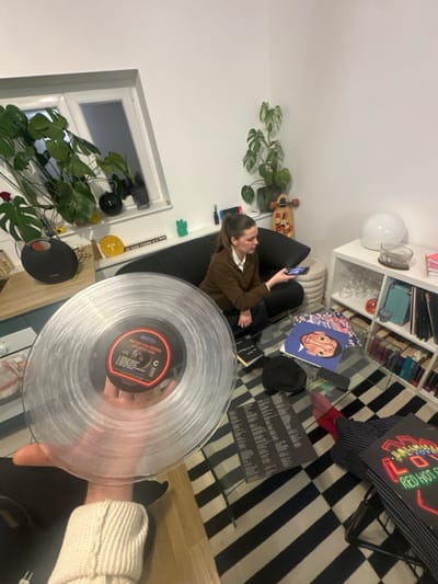 A person reclines on a dark grey sofa, using their phone. A clear vinyl record is held up in the foreground. A glass coffee table displays several vinyl album covers including Mac Miller and Red Hot Chili Peppers. A bookshelf is visible behind the sofa with various items. A window with plants sits to the left. The floor has a black and white striped rug.
