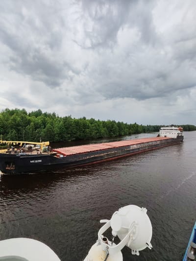 This media file captures a scenic view of a barge on a river, with a lush green forest lining the banks. The barge, identified by the Cyrillic text "МЕДОН" on its hull, is loaded with what appears to be cargo covered by red tarpaulins. In the foreground, part of another vessel is visible, featuring a white circular light fixture. The river's surface is dark and slightly rippled, suggesting a calm waterway. The sky is overcast with heavy, grey clouds, indicating a cloudy or potentially rainy day. The overall setting is that of a natural, tranquil environment, likely a river in a rural or forested area. The time of day is not explicitly indicated but appears to be daylight, given the visibility. The mood is serene and observational, focusing on the barge and its surroundings. The location is identified as Novokemskii, Russia.
