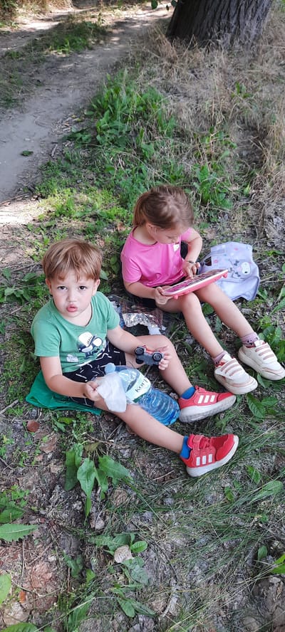 In Vadul lui Vodă, Moldova, two young children are seated on the ground amidst grass and dirt. A boy, wearing a green t-shirt and patterned shorts, sits on a green mat. He holds a toy car and a bottle of water with a white cloth. Beside him, a girl in a pink t-shirt is engaged with a pink tablet. A grey backpack with a dog illustration rests behind her. The environment appears to be outdoors, possibly near a path or wooded area, with a large tree trunk visible in the background.