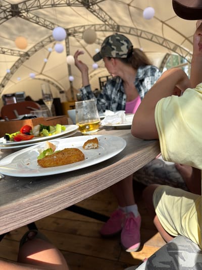 The image shows a group of people sitting at a wooden table in an outdoor setting. The scene appears to be a casual dining experience, possibly at a restaurant or cafe, with a large, tent-like structure overhead supported by metal beams. Several white and light brown spheres are hanging from the ceiling of the tent, some of which are illuminated.

The foreground features plates of food on the table. One plate contains a salad with lettuce, tomatoes, and other vegetables, while another has a breaded and fried food item, possibly a fish stick or chicken finger, with a piece of bread and lettuce. A glass of amber liquid, likely beer or juice, is also on the table.

In the background, a person wearing a camouflage hat and a plaid shirt is reaching out, possibly interacting with one of the hanging spheres. Another person is visible wearing a yellow t-shirt and pink sneakers. A third person is seen wearing a brown hat and a grey and white patterned garment.

The lighting suggests it is daytime, with sunlight casting shadows. The overall atmosphere seems relaxed and informal. The location is identified as Morozovo, Russia. There is no visible text in the image.