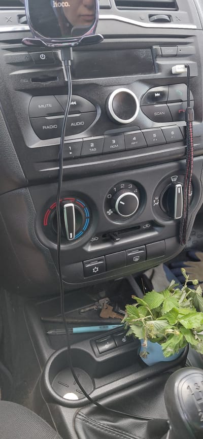 The image displays the interior of a car, specifically the dashboard area. In the foreground, a gear shift lever is visible on the right. A cup holder with a circular rubber insert is located in the center console, partially obscured by a black cable. To the left of the cup holder, keys and a blue pen are placed. A small potted plant with green leaves sits to the right of the keys. The dashboard itself features a car stereo system with various buttons and knobs, including those for radio, audio, and climate control. A phone mount with a "hoco." logo is attached to the top of the dashboard, holding a phone that reflects a person's face. The overall impression is a functional, everyday car interior, likely in a private vehicle.
The provided image does not contain any specific visual cues that would allow for the precise determination of its location within Agapovka, Russia, or any other specific geographic area. The scene depicts a generic car interior.