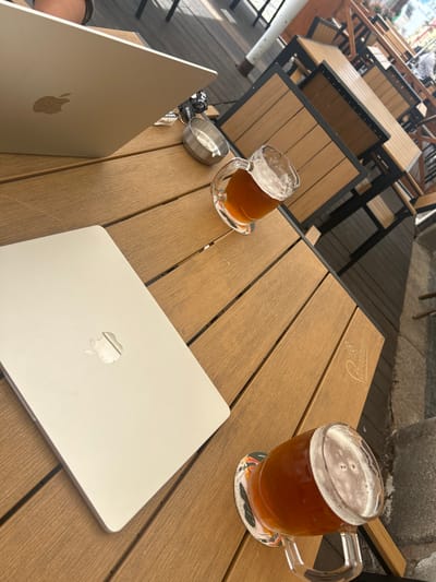 This high-angle shot captures an outdoor cafe setting, likely in Liberec, Czechia, during bright daylight. The primary subjects are a light brown slatted wooden table, two Apple MacBooks, two glass beer mugs filled with a golden-brown beverage topped with foam, a metal ashtray, and a set of keys.
The table features a faint, embossed "Staropramen" logo, a Czech beer brand, which is also visibly printed on both beer mugs. One mug is nearly full and rests on a colorful coaster with indistinct graphics, while the other is about half-full.
Two silver-colored MacBooks are open on the table, indicating ongoing use. One laptop's screen is slightly visible, while the other's lid faces up, prominently displaying the Apple logo. Next to the laptop on the left, a set of keys with a lanyard partially showing the word "work" is placed next to a small, circular metal ashtray.
In the background, out-of-focus wooden tables with dark metal frames and chairs suggest a larger outdoor dining or seating area. The ground beyond the table appears to be a paved surface. The scene conveys a relaxed atmosphere, potentially for remote work or leisure, enjoying a beer outdoors.