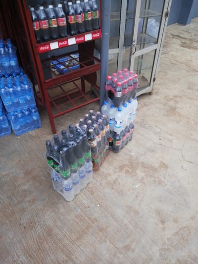 This image depicts an outdoor scene in Ajasse Ipo, Nigeria, likely a roadside stall or small shop. The primary subjects are stacks of bottled beverages, predominantly Coca-Cola and water. Several racks, one red and metal, another a glass display cabinet, are visible, holding more bottles. The ground is a textured concrete surface.

In the foreground, on the right, are several plastic-wrapped cases of dark-colored bottles, possibly Coca-Cola or a similar beverage, with some lighter-colored bottles on the bottom layer. To the left of these, and further back, are numerous clear plastic bottles of water, also plastic-wrapped. The red metal rack, visible on the left side of the image, is filled with more Coca-Cola bottles. The glass display cabinet, on the right, is mostly empty with shelves visible.

The lighting suggests it is daytime, with no strong shadows indicating a specific time of day. The overall impression is one of a commercial setting where drinks are sold. Text visible includes "Coca-Cola" on several bottles and signage, and "ALKA" and "AQUAVI" on the water bottles. The scene is static, with no people or discernible activity beyond the display of goods.