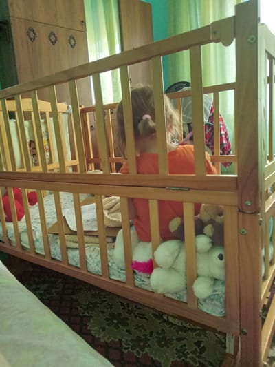 The image depicts an indoor scene within a room, likely a child's bedroom, located in Kitskany, Moldova. The primary focus is a wooden crib filled with stuffed animals and a child. The child, seen from behind, wears an orange shirt and has blonde hair tied in a ponytail. Various plush toys, including what appear to be teddy bears and a white bunny, are scattered within the crib. The crib has wooden railings and a mattress covered with a patterned sheet. In the background, a wooden wardrobe with decorative elements is visible, along with green curtains and a hint of a turquoise wall. The floor is covered by a patterned rug. The overall lighting suggests a moderately lit environment.