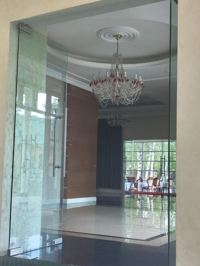 The media file shows a reflection in a glass enclosure. In the reflection, a chandelier with crystal prisms and red accents hangs from a white ceiling with ornate molding. Beyond the chandelier, a window reveals a lush green outdoor scene with trees. Inside the reflected space, there is a table and chairs, suggesting a dining or seating area. The interior also features dark fabric drapes and what appears to be a wall with vertical wooden panels. The foreground of the media file is dominated by the glass enclosure itself, with a metal railing and supporting fixtures. The glass has a slightly textured or frosted lower section. The overall impression is of an indoor space with a view of nature. The lighting suggests it could be daytime, with natural light filtering through the window. No people or specific activities are directly visible, only the static elements of the reflected environment and the glass structure.