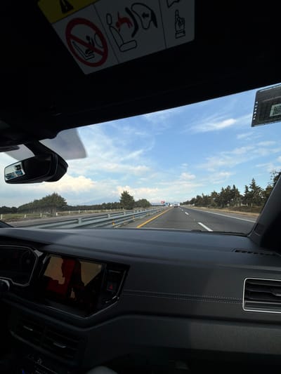 An interior car view showcases a highway stretching into the distance under a blue sky with scattered clouds. A guardrail lines the road's edge, bordered by trees. The dashboard is visible, with a screen displaying a map. A rear-view mirror and sun visor with warning labels are also in view. The image is rotated, giving a tilted perspective.