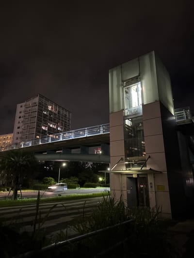 This image captures a nighttime scene in Chiba, Japan, focusing on a modern building with an illuminated elevator shaft and a connecting walkway. The overall mood is one of quiet urban life after dark.

The primary subject is a multi-story building, its facade featuring a combination of opaque panels and large glass sections that reveal the inner workings of the elevator. The elevator shaft itself is brightly lit, showcasing the mechanical components and the empty car within. A transparent, illuminated walkway extends from the upper level of this building, suggesting an elevated pedestrian path or a connection to another structure.

In the background, another residential building is visible, its numerous windows lit from within, indicating occupancy. The scene is set against a dark, cloudy night sky. A street runs alongside the buildings, with a single streetlight casting a pool of light on the pavement and grass verge. The presence of trees and dense foliage on the right side of the frame adds to the natural elements within the urban landscape.

A car is seen driving on the road in the distance, its headlights creating streaks of light due to the long exposure or movement. This adds a dynamic element to the otherwise static architecture.

There is some visible text on the building, including what appears to be a yellow caution sign and some smaller icons or labels near the elevator doors. However, the text is too small and indistinct to be fully deciphered. The scene conveys a sense of order and functionality, typical of a modern Japanese urban environment, with a hint of isolation due to the nighttime setting and the absence of people.