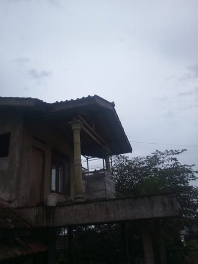 The image depicts the exterior of a concrete building in Tha Yet Gwa, Myanmar. The building has a dark tiled roof that overhangs a balcony supported by concrete beams and a single ornate pillar. A door and window are visible on the facade, and a white sack sits near the window on the balcony. Trees are visible in the background against an overcast sky.