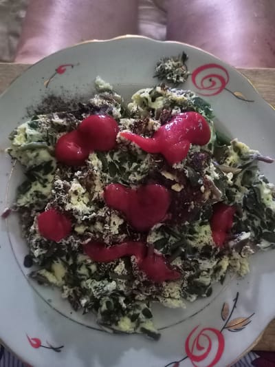 The image shows a close-up, overhead view of a serving of scrambled eggs with chopped greens, topped with several dollops of a red sauce or paste, possibly chili paste or a tomato-based relish. The dish is served on a white plate with a decorative floral pattern in red and gold. The plate is positioned on a wooden surface, and in the background, a person's legs are visible, suggesting the meal is being consumed. There is no indication of the city of Ryabichev, Russia, or any other specific geographical location within the image itself. The scene depicts a simple meal prepared and presented for consumption.