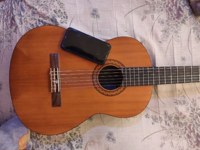 This image depicts a classical guitar and a smartphone resting on a floral-patterned fabric surface in Staufenberg, Germany.
The primary object is a light brown, wooden classical guitar, oriented with its headstock out of frame to the left and its body extending towards the right. It features a circular soundhole adorned with a rosette design. Six strings are visible, running from the bridge (a dark wooden piece on the lower body) across the soundhole and a dark fretted fingerboard.
A dark-screened smartphone, encased with a visible blue edge, is positioned diagonally across the upper right quadrant of the guitar's body, partially obscuring the upper bout and part of the soundhole area. The phone's screen is dark and reflective.
The guitar and phone rest on a light-colored fabric surface, possibly a bedspread or blanket, which displays a repeating pattern of stylized flowers and leaves in shades of purple, blue, and green. The fabric is slightly creased. No individuals or other significant objects are visible in the frame.