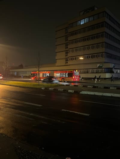 The image captures an urban street scene at night in Schweinfurt, Germany, characterized by dim lighting and wet surfaces, suggesting recent rain.

In the foreground and midground, a tall, multi-story building dominates the left side. The building appears to be an office or commercial structure, with numerous rectangular windows across its facade. The logo "AOK" is prominently displayed on the building in at least two visible locations, indicating it houses an AOK health insurance office. A single, brightly lit window on the lower floors stands out against the darker building.

Along the street next to the building, a long, red articulated bus is visible. The bus displays the route number "5" on its front and side. Visible German text on the side of the bus includes phrases like "Wir versorgen Sie - rund um die Uhr" (We supply you - around the clock) and "Service-Bus für... 15.000 €," likely part of an advertisement. Several individuals are standing on the sidewalk near the bus and the building, with some figures appearing blurry due to motion or distance.

The street itself, occupying the right side of the frame, is dark and reflective from the moisture, mirroring the glow of streetlights and vehicle headlights. Road markings, including white lines and a striped curb, are visible. A narrow strip of grass with a bare tree separates the sidewalk from the road. In the distance, other blurry vehicles suggest ongoing traffic. The overall impression is a typical city night scene, quiet yet active with public transport and pedestrians.