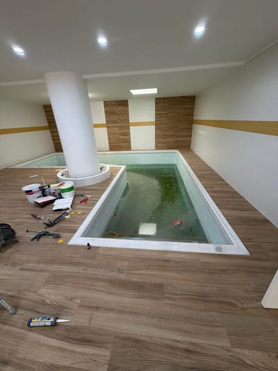 The image depicts an indoor scene, likely a bathroom or spa area, featuring a small swimming pool or jacuzzi filled with greenish water. The pool is surrounded by tiled walls and a wooden-effect floor. Several construction or maintenance tools are scattered on the floor around the pool, including spools of tape, a caulking gun, and other small items. A large, white, cylindrical object, possibly a pillar or decorative element, stands near the pool. The ceiling features recessed lighting and decorative yellow and white stripes. The overall impression is one of an ongoing project, perhaps the installation or renovation of a swimming area. There are no people visible in the image.