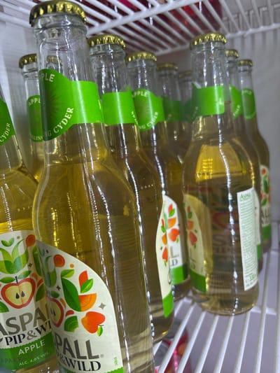 The image shows a close-up of several bottles of Aspall Apple Cyder in a cooler or refrigerator. The bottles are stacked horizontally, with the neck and caps visible on the left and the labels prominently displayed. The labels show the Aspall brand name, details about the drink being a sparkling apple cyder with 4.0% alcohol by volume, and apple-themed imagery. The cooler has white racks. The location of the photo is not provided, it cannot be determined that this is the city of Vitez, Bosnia and Herzegovina.