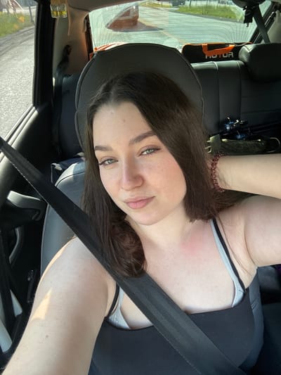 A young woman with long dark hair and green eyes is in the passenger seat of a car, taking a selfie. She is wearing a black tank top and a grey bra, with a black seatbelt crossing her chest. A reddish area is visible on her upper chest. In the background, the back seats of the car are visible, along with a bag with the word "MOTOR" on it and what appears to be fishing rods. The exterior of the car, seen through the windows, suggests an outdoor setting with a road and some greenery, possibly on the outskirts of Pesterevo, Russia.