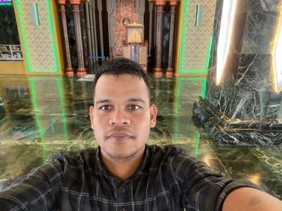 The image is a selfie taken from a low angle, showing a man in the foreground. He is wearing a black and white plaid shirt and has dark hair and brown eyes. His expression is neutral. The background suggests an indoor setting with elaborate architectural features. To the left, there are decorative pillars with copper accents and a patterned wall illuminated by green lighting. In the center, a highly ornate wooden structure with intricate carvings and golden accents is visible. To the right, a dark, veined marble structure is accented by bright, vertical white and yellow lighting. The floor appears to be polished green marble, reflecting the ambient light and the surroundings. The overall ambiance suggests a place of worship or a grand public building, possibly within a complex such as the Baiturrahman Grand Mosque in Banda Aceh, which is located in the province of Aceh, Indonesia. Aluekambuk is a village in the Aceh Besar Regency, which is part of the province of Aceh, Indonesia, and it is plausible that such a structure could be found in or near a significant religious or public site within this region.