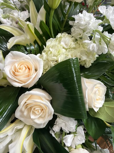 A close-up, overhead shot captures a vibrant bouquet of predominantly white and cream flowers. The arrangement features several creamy white roses with tightly furled centers, interspersed with delicate white lilies, white hydrangeas with ruffled petals, and smaller white filler flowers. Lush, dark green leaves, some glossy and others matte, provide a contrasting backdrop. A prominent, dark green, glossy leaf curves across the center of the frame, adding a sculptural element. The lighting suggests an indoor setting, possibly a florist's shop or a home, with no discernible background elements to indicate a specific location within Cumming, Georgia, United States.