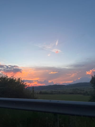 The image presents a landscape scene under a colorful sunset sky. The sky transitions from a clear light blue on the left to a fiery mix of oranges, reds, and pinks towards the center and right. These vibrant colors illuminate the clouds, some of which are layered and textured. A row of dark green hills or mountains is visible in the distance. In the foreground, there are trees and vegetation, appearing dark against the bright sky. A blurred metallic structure is also visible on the right side of the image, partially obscuring the view. The location is presumably near Ponor, Bulgaria, based on the prompt.