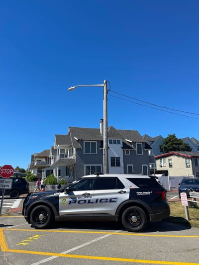 A police SUV is parked on a paved road, with yellow lines painted on the asphalt, in front of a row of houses and a white picket fence. The SUV is black and white, with "OAK BLUFFS POLICE" written on its side, along with a shield emblem. The SUV's doors are closed, and it appears to be stationary. Behind the SUV, several people are visible walking on a sidewalk, and further back, houses with shingled exteriors and white trim are present. A stop sign with a "No Right Turn" indicator is visible to the left of the police SUV. Overhead, a street light on a wooden pole is present, with wires strung between poles against a clear blue sky. A sign indicating "POLICE PARKING OSCAR 3 VIOLATORS WILL BE TOWED" is posted on the right side of the image. The environment appears to be a sunny day in a residential area.