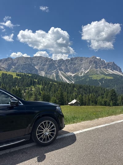 The image captures a scenic mountain landscape with a car parked on the side of a road. The mountain range dominates the background, with rugged peaks and slopes covered in lush green forests. Patches of snow are visible on the higher elevations, suggesting a cooler climate even in warmer months. A small wooden structure, possibly a shed or a small cabin, is nestled amongst the trees, adding a touch of rustic charm to the scene. The sky is a vibrant blue with scattered white cumulus clouds, indicating fair weather and a clear day.
In the foreground, a portion of a dark-colored car is visible, with its wheel resting on the asphalt road. The car appears to be parked on the shoulder, next to a grassy verge. The road has a white line marking its edge. The perspective of the image suggests the viewer is looking from the car outwards towards the landscape, or perhaps from a similar vantage point next to the car.
The time of day appears to be daytime, likely mid-morning or afternoon, given the bright sunlight casting shadows. The overall mood is serene and picturesque, evoking a sense of tranquility and appreciation for nature's grandeur. The location context of San Martin de Tor - San Martino in Badia - St. Martin in Thurn, Italy, places this scene in the Dolomites, a renowned mountain range in the Alps known for its dramatic peaks and natural beauty. No visible text is present in the image.