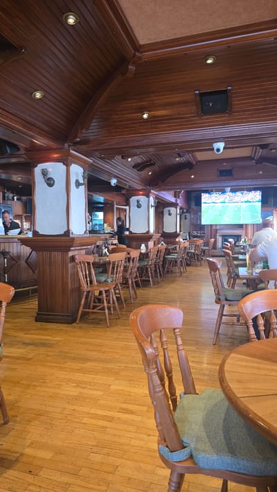 The image shows the interior of a bar or restaurant with a traditional wooden decor. The scene is illuminated by warm lighting and appears to be during the daytime, as evidenced by the bright television screen showing a soccer match. The bar area is lined with wooden chairs, some of which are stacked. A few people are visible: one person in a white shirt stands near the bar, and another person in a black uniform is behind the bar, seemingly working. A security camera is mounted on the wooden wall. The floor is made of polished wooden planks. The overall atmosphere seems calm and perhaps preparing for the day's business, given the stacked chairs and subdued activity. The location cues, such as the style of the interior and the mention of Parekklisia Community, Cyprus, suggest a Mediterranean setting.