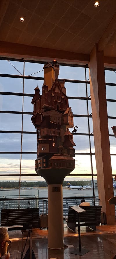 This image captures a view from inside Stockholm-Arlanda Airport, likely within a lounge area, overlooking the tarmac. A prominent, multi-layered sculpture resembling a whimsical, stacked village or castle dominates the center foreground, silhouetted against a large window. The sculpture is made of wood and features numerous windows, roofs, and architectural details. A red light is visible at the very top of the sculpture.
Through the expansive window, the airport tarmac is visible, with a Ryanair airplane taxiing or parked in the distance. The sky is a muted twilight, with hints of sunset colors. In the foreground, seating benches with metal slats are visible, along with a few pieces of luggage, including a silver suitcase and a black carry-on bag. A man wearing glasses is partially visible on the left, and another person is seated on a bench on the right, facing away from the camera. The overall environment suggests an indoor waiting area at an airport, with natural light streaming in from the large windows.