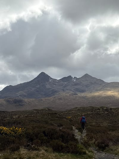 The image presents a mountain landscape on the Isle of Skye in the United Kingdom under an overcast sky. A prominent mountain with a rocky peak dominates the left side of the frame, its slopes exhibiting barren terrain with patches of snow near the summit. To the right, a hillside descends, covered in a mix of grasses and scrub vegetation. A hiker with a blue backpack and red jacket is seen on a trail traversing the hillside. Yellow flowering shrubs are visible along the edge of the hillside. The overall atmosphere is remote and rugged.