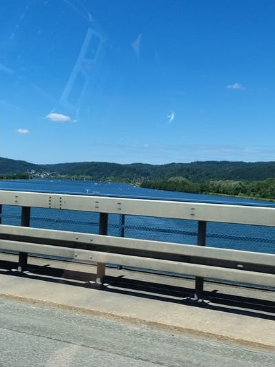 This is a vertical, medium-angle, eye-level, outdoor shot taken from a bridge in Barbing, Germany. The scene depicts a wide, blue river with forested hills on the opposite bank. Several small, white buoys are visible in the water. The bridge railing, made of grey metal bars and posts, runs along the foreground. The sky is a clear, bright blue with two small, white clouds. The image has a slight reflection on the upper left side. The foreground also shows a portion of the asphalt road surface of the bridge.