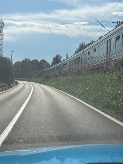 The image depicts a curving asphalt road with a white line dividing the lanes, leading through a rural landscape. To the right of the road, tall green grass grows alongside a train track where a long train with multiple carriages is parked. The train appears to be older, with visible windows and a faded exterior. In the background, dense green trees form a treeline. Above, a bright blue sky is dotted with fluffy white clouds. On the left side of the road, a tall cellular tower stands prominently, and a street light is visible behind it. A concrete barrier with a metal railing runs along the left edge of the road. The image appears to have been taken from a moving vehicle, as suggested by the blue hood or dashboard in the foreground. The location is stated to be in the city of Bistrice, Montenegro.